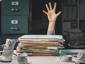 Hand hinter einem Berg von Papieren auf dem Schreibtisch