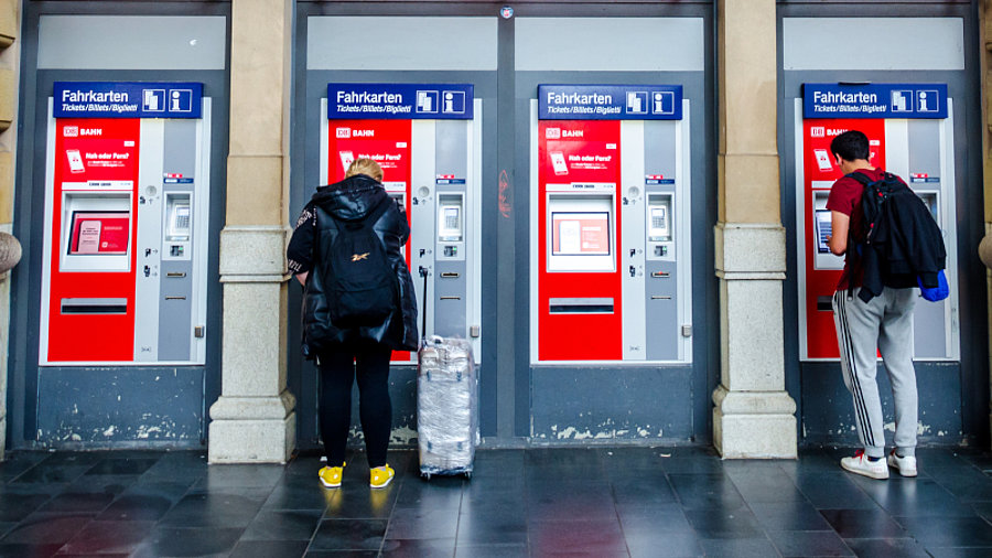 Zwei Personen stehen vor Fahrkartenautomaten im Bahnhofseingang