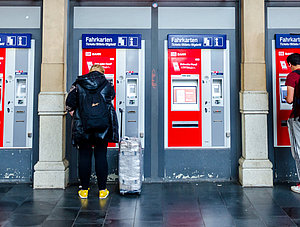 Zwei Personen stehen vor Fahrkartenautomaten im Bahnhofseingang