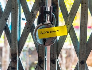 Ein Vorhängeschloss, das an einem geschlossenen Gittertor befestigt ist. Am Schloss hängt ein gelber Zettel mit der Aufschrift "Lockdown". Im Hintergrund sind verschwommene Bäume und ein sonniger Außenbereich zu sehen.