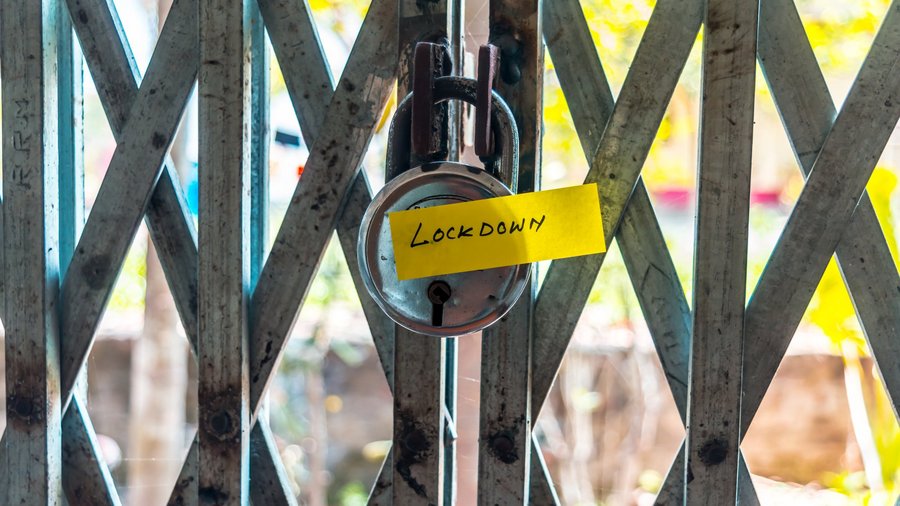 Ein Vorhängeschloss, das an einem geschlossenen Gittertor befestigt ist. Am Schloss hängt ein gelber Zettel mit der Aufschrift "Lockdown". Im Hintergrund sind verschwommene Bäume und ein sonniger Außenbereich zu sehen.