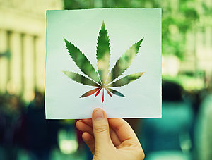 Hand, die ein Papierblatt mit einem Symbol f&uuml;r Marihuana-Bl&auml;tter auf einem &uuml;berf&uuml;llten Stra&szlig;enhintergrund h&auml;lt.