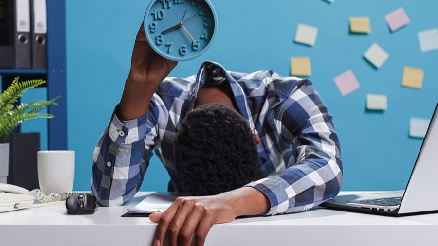 &Uuml;berstunden: B&uuml;ro-Angestellter mit Kopf auf Tisch und Uhr in der Hand