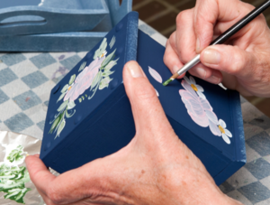 Eine Nahaufnahme zeigt eine Person, die eine dunkelblaue Holzbox mit einem Pinsel und Acrylfarben bemalt. Die H&auml;nde der Person sind sorgf&auml;ltig dabei, ein florales Design mit rosa und wei&szlig;en Bl&uuml;ten sowie gr&uuml;nen Bl&auml;ttern auf die Oberfl&auml;che der Box zu malen. Im Hintergrund befinden sich weitere bemalte Holzgegenst&auml;nde sowie verschiedene Farbt&ouml;pfe und eine Farbpalette mit verschiedenen Farbmischungen. Die Person tr&auml;gt ein wei&szlig;es Oberteil und ein silbernes Armband.