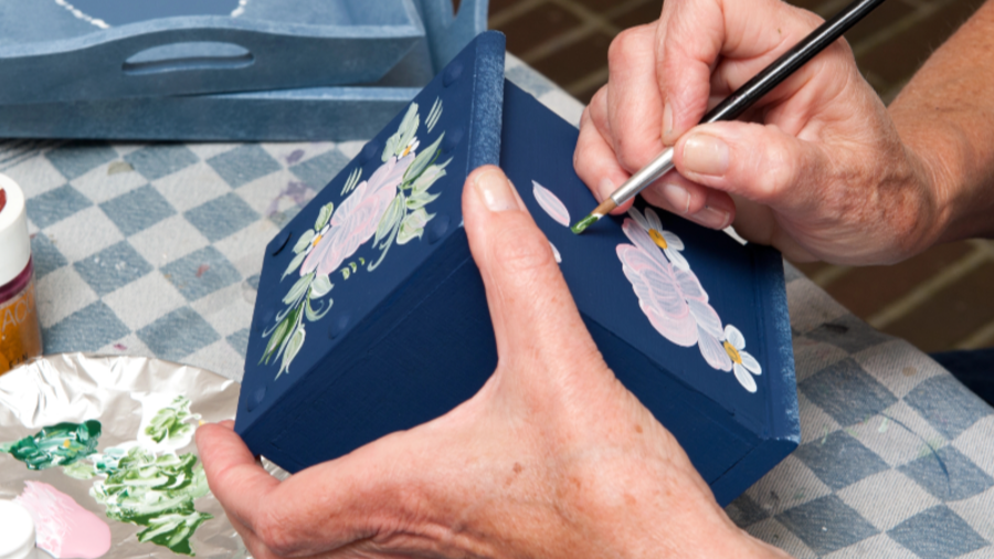 Eine Nahaufnahme zeigt eine Person, die eine dunkelblaue Holzbox mit einem Pinsel und Acrylfarben bemalt. Die H&auml;nde der Person sind sorgf&auml;ltig dabei, ein florales Design mit rosa und wei&szlig;en Bl&uuml;ten sowie gr&uuml;nen Bl&auml;ttern auf die Oberfl&auml;che der Box zu malen. Im Hintergrund befinden sich weitere bemalte Holzgegenst&auml;nde sowie verschiedene Farbt&ouml;pfe und eine Farbpalette mit verschiedenen Farbmischungen. Die Person tr&auml;gt ein wei&szlig;es Oberteil und ein silbernes Armband.