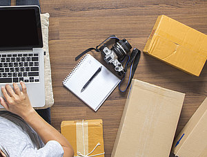 Draufsicht von Person, die neben Kartons auf dem Boden sitzt und mit dem Notebook auf dem Scho&szlig; shoppt