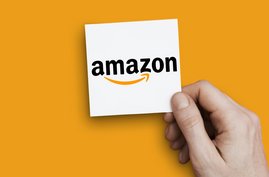 Eine Hand hält einen Zettel mit dem Amazon-Logo vor gelben Hintergrund