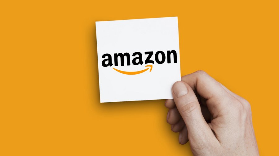 Eine Hand hält einen Zettel mit dem Amazon-Logo vor gelben Hintergrund