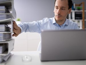 Mann schiebt Aktenordner weg und arbeitet mit Laptop.