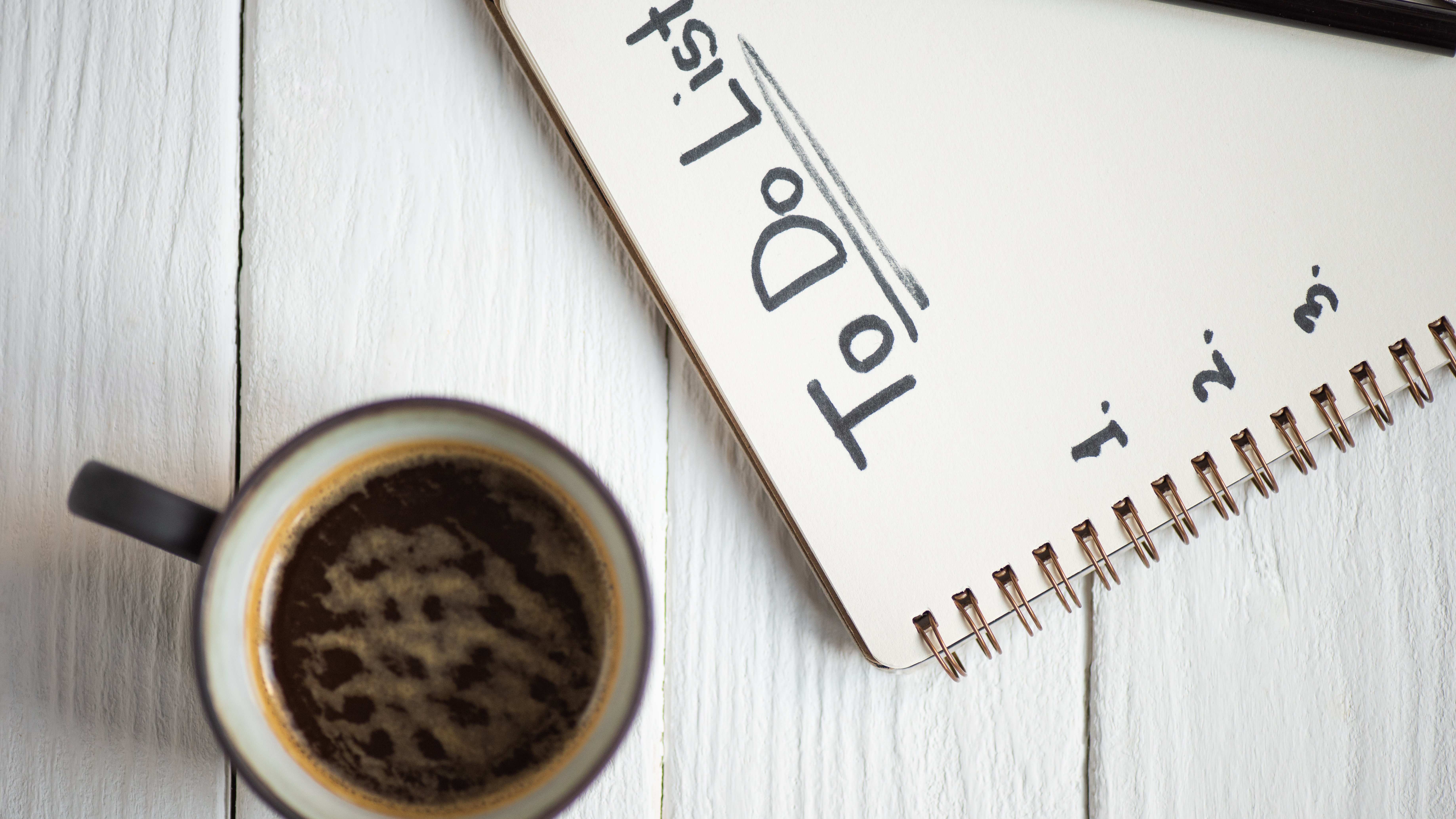  Das Bild zeigt einen Notizblock mit der Aufschrift "To Do" und eine Tasse Kaffee auf einem weißen Holztisch. Der Notizblock ist aufgeschlagen und ein Stift liegt daneben.