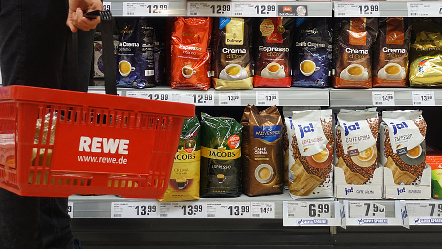 K&auml;ufer geht mit Rewe-Einkaufskorb an Regal vorbei und packt Produkt ein.