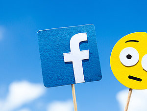 Facebook-Logo und erstaunter Smiley an Holzstab vor blauem Himmel.