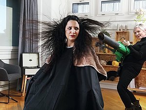 Frau mit abstehenden Haaren und Friseurin mit Laubbläser